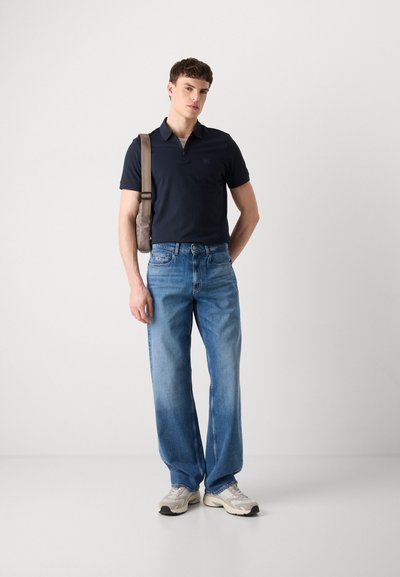 Polo-shirt marine à manches courtes et zip, associé à un jean droit bleu clair et des baskets beiges ; mannequin portant un sac à bandoulière gris.