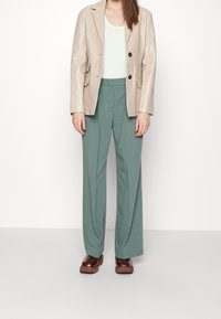 Veste en cuir beige avec deux boutons, pantalon large vert, et chaussures à plateforme marron ; textures lisses et couleurs neutres.