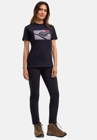 Zwart T-shirt met een grafisch ontwerp van een voet en geometrische vormen, gecombineerd met zwarte broeken en bruine wandelschoenen.