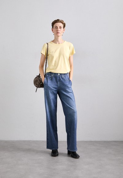 Gul kortermet t-skjorte, lyse denim vide bukser og svarte ankelstøvler. Modellen holder en liten brun veske. Nøytral bakgrunn.