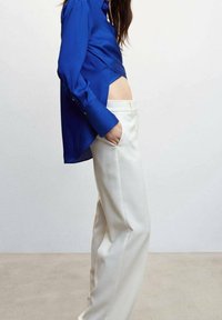 Chemise en satin bleu à manches longues et ourlet arrondi, associée à un pantalon large blanc. Les textures lisses et les lignes épurées sont très présentes.