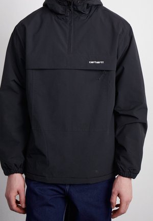 Czarna bluza Carhartt z kapturem, zapinana na zamek z przodu, z kieszenią na klatce piersiowej na zamek, noszona z ciemnoniebieskimi dżinsami.