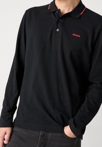 Schwarz langärmliges Polohemd aus Baumwolle mit roten Akzenten am Kragen und einem kleinen roten "HUGO"-Logo auf der Brust.