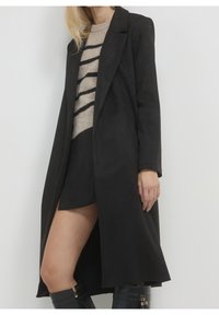 Femme portant un long manteau noir, un pull beige à rayures noires, une jupe noire et des bottes noires montantes, debout devant un fond blanc uni.