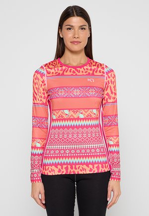 Frau trägt ein langärmeliges, eng anliegendes pinkfarbenes Oberteil mit mehrfarbigen geometrischen und Tiermustern, kombiniert mit schwarzen Hosen.