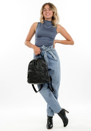 Smaidoša sieviete zilā bezrūtainā krekliņā un augsta vidukļa džinsos krusto kājas, vienā rokā tur melnu ādas mugursomu, ar otru roku balstās uz gurna.