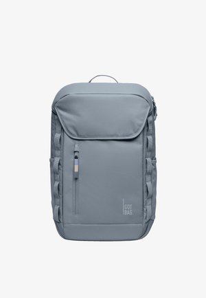 Mochila de tela gris con solapa frontal, correas laterales, bolsillo con cremallera y detalle de logo. Textura suave y forma rectangular.