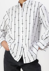 Chemise blanche à col avec un motif répété de logo noir, fermeture à boutons et manches longues. Portée avec un jean foncé.