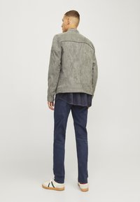 Grey suede jacket with a high collar, back yoke detail, paired with dark blue jeans and white sneakers, standing against a plain background.