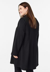 Žena s dlouhými hnědými vlasy, která má na sobě stříbrné náušnice a volně padnoucí černý kabát, se mírně otáčí a usmívá proti bílému pozadí.