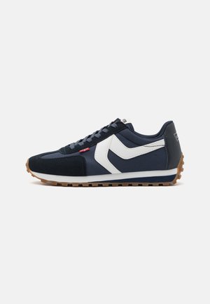 Navyblaue Sneakers mit einem weißen V-förmigen Akzent, Obermaterial aus Wildleder und Textil, Gummisohle und grauen Schnürsenkeln. Auffälliges rotes Logo-Etikett an der Seite.