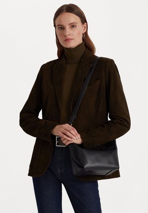 Kvinde iført mørkebrun rullekrave og blazer, der holder en sort læder skuldertaske mod en ensfarvet baggrund.