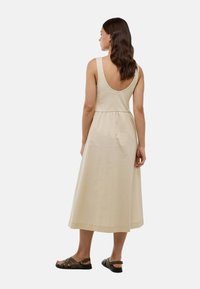 Robe maxi sans manches beige avec un dos en scoop, taille cintrée et jupe fluide. Portée avec des sandales à lanières sombres. Texture en coton lisse.