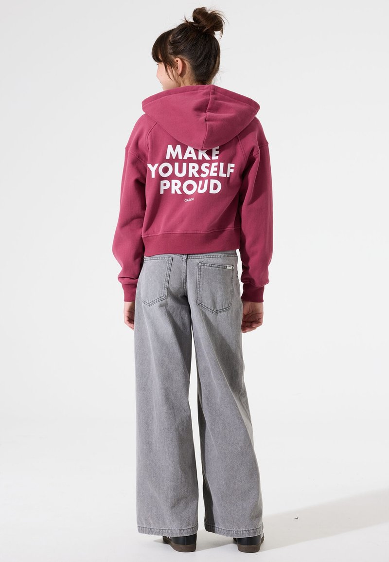 Sudadera corta burdeos con el texto blanco "HAZTE ORGULLOSO" en la parte posterior; combinada con jeans gris holgados y botas negras de suela gruesa.