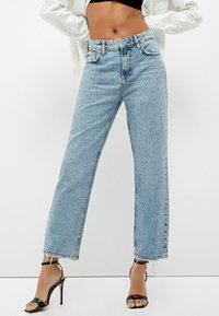 Jeans de mezclilla azul claro con un corte recto, con un dobladillo deshilachado y salpicaduras oscuras. Combinados con sandalias negras de tacón alto.