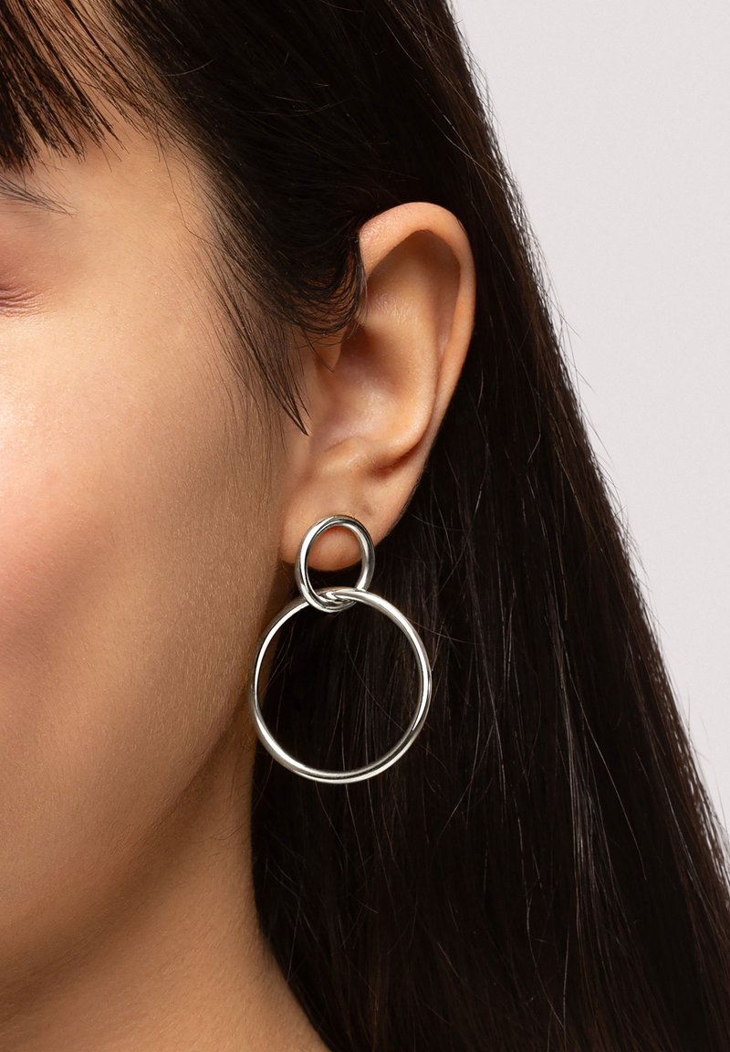 Pendientes de aro de plata que presentan formas circulares interconectadas, con una textura metálica suave y un acabado pulido, colgando cerca de la oreja.