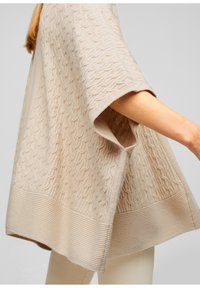 Poncho in maglia beige, con un motivo a onde testurizzato e un orlo a costine. Vestibilità ampia con maniche larghe, realizzato in materiale morbido e leggero.