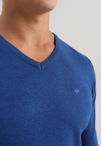 Blå V-ringad tröja som är tillverkad av mjukt material, med en subtil texturerad yta och en liten logotyp på vänster bröstområde.