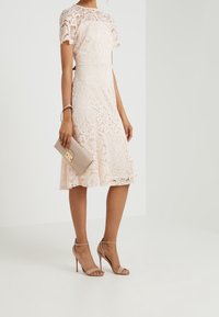 Žena oblečená v růžové krajkové koktejlové šaty s délkou ke kolenům, se světle hnědými vysokými sandály a držící béžovou kabelku s zlatým zapínáním.