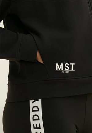 Sweat-shirt noir avec main dans la poche latérale et texte "MST FREDDY" au-dessus de l'ourlet ; leggings noirs avec bande verticale blanche portant le texte "FREDDY".