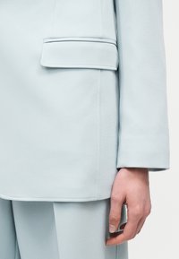 Κοντινό πλάνο ενός ανοιχτόχρωμου γαλάζιου ραμμένου κατά παραγγελία σακακιού με λεπτομέρεια τσέπης με καπάκι και ταιριαστό παντελόνι, που δείχνει ένα χαλαρό χέρι στο πλάι.