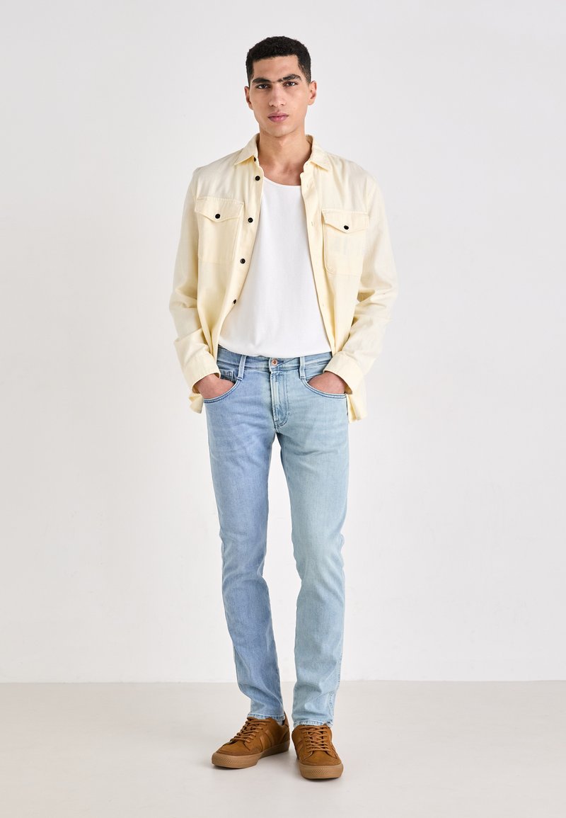 Lichtgele overhemden, witte tanktop, lichtblauwe jeans en bruine sneakers. Het overhemd heeft twee borstzakken en zwarte knopen.