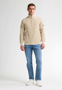 Beige ribbad tröja med halvlång dragkedja och långa ärmar, kombinerad med ljusblå jeans och vita sneakers, vilket skapar en avslappnad outfit.