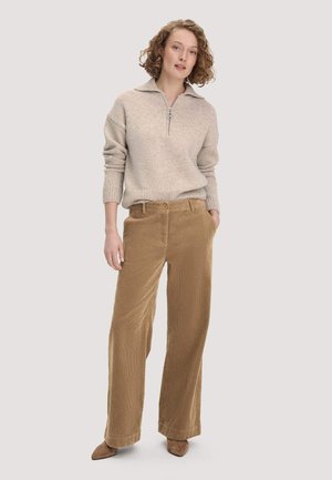 Vrouw met krullend haar die een beige trui met rits en brede bruine corduroy broek draagt, staat met haar handen in de zakken.