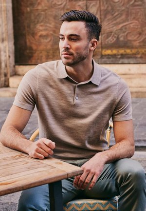 Mann mit beigem Poloshirt sitzt draußen an einem Holztisch und blickt mit neutralem Ausdruck zur Seite.