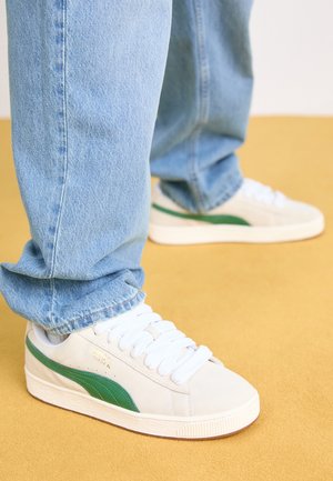 Persona con vaqueros azul claro y zapatillas blancas con una franja verde, de pie sobre un suelo de color mostaza.