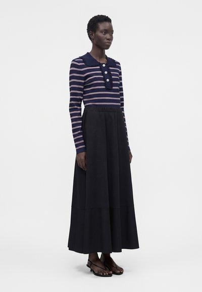 Γυναίκα που φοράει μακρυμάνικη μπλούζα με ναυτικό μπλε και ροζ ρίγες, με μεγάλα κουμπιά, και μαύρη φούστα μέχρι τον αστράγαλο, στέκεται με τα χέρια στα πλευρά.