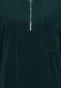 Teal corduroy top met een vooraanstaande rits en een borstzak. De verticale ribbelstructuur verbetert de duurzaamheid en visuele aantrekkingskracht.