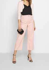 Ljusrosa korta vida jeans som är kombinerade med en svart topp med spetsdetaljer och en blekrosa kofta med vågkant; accessoarer med en svart väska.