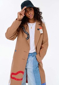 Žena s kudrnatými vlasy, oblečená v hnědém kabátu s popruhy "NESTAŇ SE", světle modrých džínách, bílém tričku, černé kšiltovce a barevných perlových náhrdelnících.