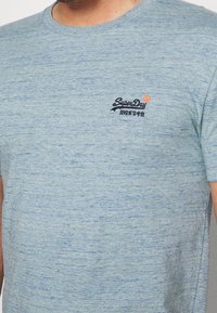 Camiseta de manga corta azul claro hecha de una mezcla de algodón texturizado, con un pequeño logo bordado de "SuperDry" en el pecho.