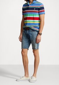 Blå randig polo med röda, gröna och gula accenter; slitna denimshorts; och vita sneakers. Läderbälte och klocka synliga.