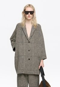 Persona vistiendo un traje de cuadros blanco y negro de gran tamaño con gafas de sol anchas, sosteniendo una bolsa marrón con una correa negra sobre un fondo blanco.