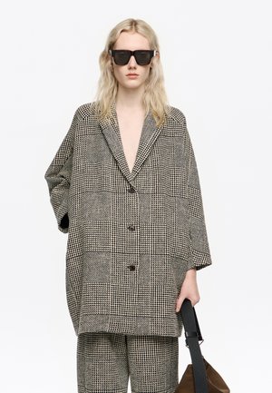 Persona vistiendo un traje de cuadros blanco y negro de gran tamaño con gafas de sol anchas, sosteniendo una bolsa marrón con una correa negra sobre un fondo blanco.