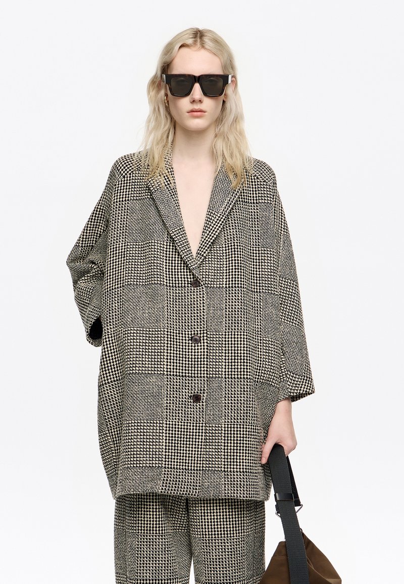 Persona vistiendo un traje de cuadros blanco y negro de gran tamaño con gafas de sol anchas, sosteniendo una bolsa marrón con una correa negra sobre un fondo blanco.