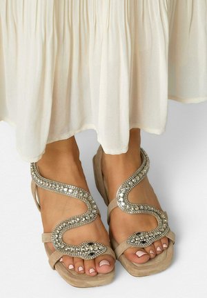 Pieds chaussés de sandales beige en forme de serpent ornées de strass argentés, associés à une longue robe blanche fluide.
