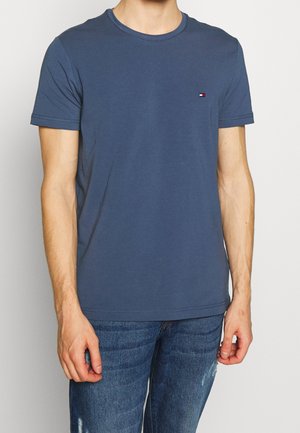 Mann trägt ein schlichtes blaues T-Shirt mit kurzen Ärmeln, einem kleinen Logo auf der Brust und leicht abgenutzte blaue Jeans.