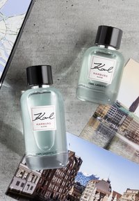Two clear glass perfume bottles with black caps, labeled "Karl Hamburg Alster," on a textured gray surface with postcards in the background.
