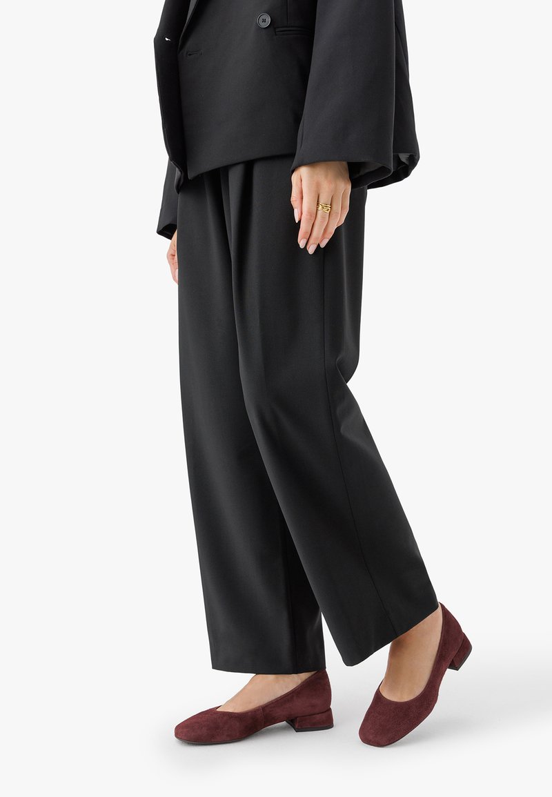 Fekete, bő szárú nadrágot és fekete blézert viselő személy, az egyik lábát kissé megemelve áll, burgundi velúrból készült, alacsony sarkú cipőt viselve.