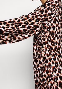 Długi rękaw odzieży w odważny leopardi wzór w czerni, brązie i beżu na białym tle, pokazany na uniesionej ręce na jednolitym tle.