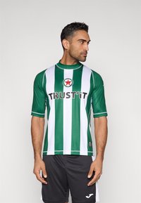 Zielono-biała koszulka sportowa w paski z krótkim rękawem, z napisem "TRUST'IT" i logo w kształcie gwiazdy. Noszona z czarnymi szortami.