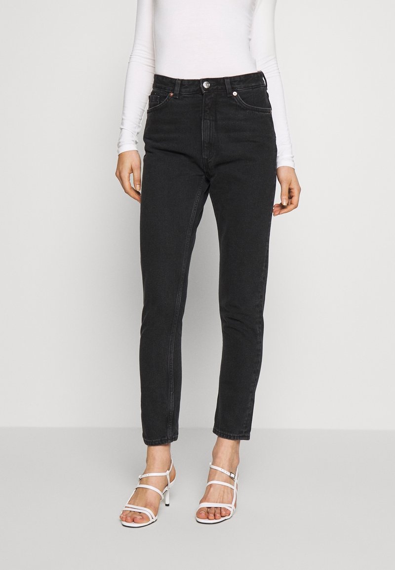 Jeans de mezclilla negra con un ajuste slim, talle alto y diseño de cinco bolsillos. Combinados con tacones blancos de tiras y una blusa ajustada de manga larga.