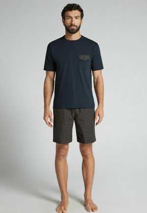 Mand står barfodet iført en sort kortærmet skjorte med ternet lomme og matchende ternede shorts på grå baggrund.