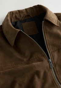 Braune Wildlederjacke mit silbernem Reißverschluss, teilweise geöffnet, zeigt das schwarze Innenfutter und den Kragen, flach auf einer weißen Oberfläche ausgebreitet.