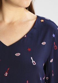 Blouse bleu marine avec un décolleté en V, ornée de motifs décoratifs d'étoiles rouges, de guitares et de lèvres de différentes couleurs sur un tissu lisse.