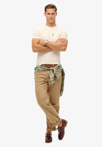 Camiseta ajustada beige, pantalones caqui con puños enrollados, camisa a cuadros verde atada a la cintura y zapatos de barco color burdeos con cordones marrones.